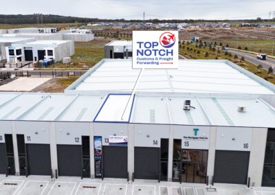 TopNotch_Site - Top Notch Customs Aerial view of a modern warehouse complex with numbered bays (10–16) and a prominent Top Notch Customs & Freight Forwarding sign on the roof. In the background, an industrial park and roads stretch across a suburban landscape.
